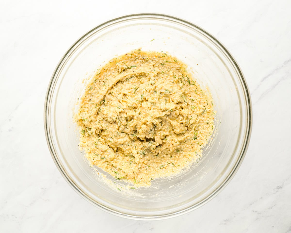 A glass bowl containing a mixture of matzo ball dough with visible flecks of herbs like dill, ready to be formed into balls.