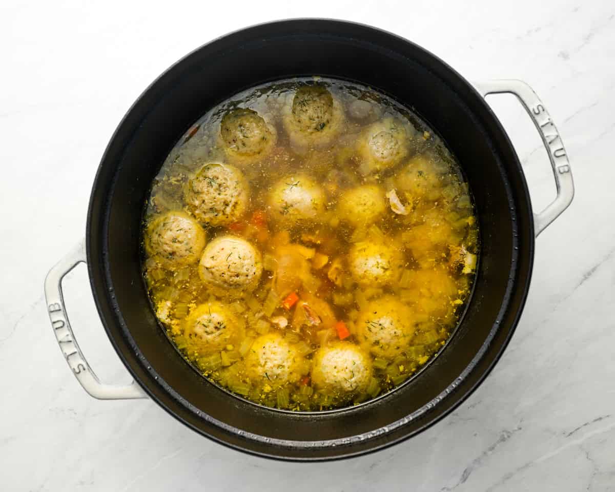 A large black pot filled with matzo ball soup, featuring matzo balls, vegetables, and a golden broth.