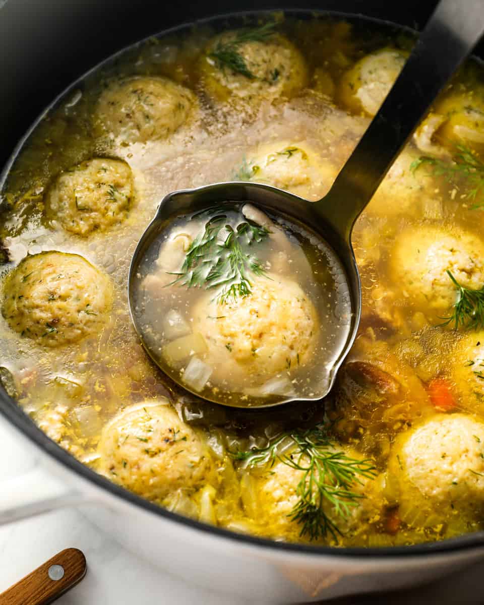 A ladle scooping matzo ball soup from a pot filled with broth, vegetables, matzo balls, and garnished with dill.