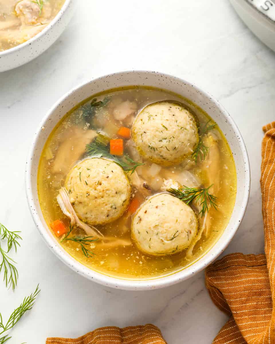 A close-up of a bowl of matzo ball soup showcasing three fluffy matzo balls in a vibrant broth with carrots and herbs.