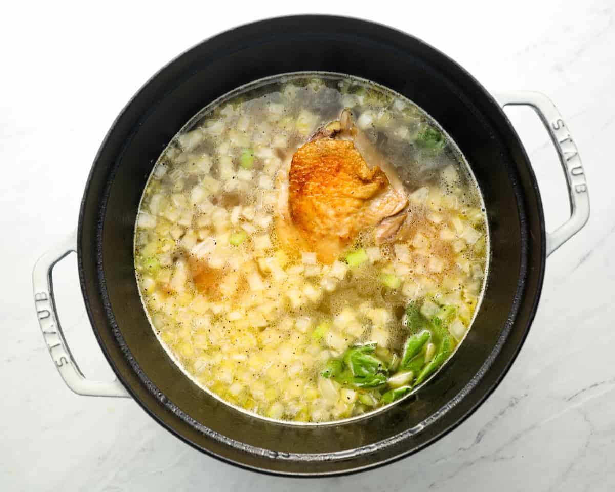 A pot filled with chicken broth, vegetables, and browned chicken thighs, simmering with herbs to create the soup.