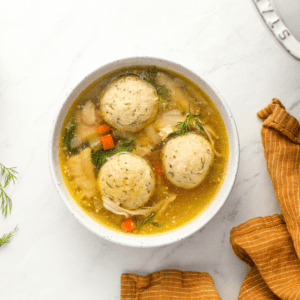 A bowl of matzo ball soup featuring three fluffy matzo balls in a golden broth with diced vegetables, shredded chicken, and fresh dill, set on a white surface with an orange-striped napkin and dill sprigs for garnish.