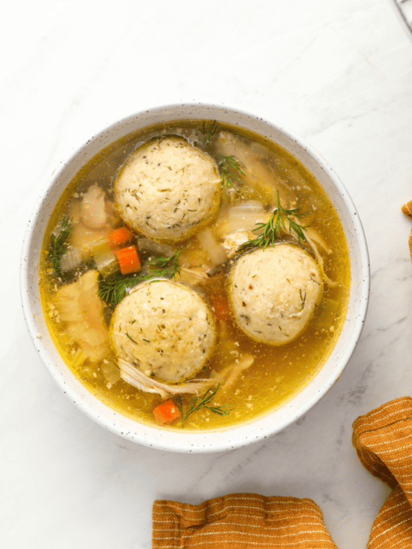 A bowl of matzo ball soup featuring three fluffy matzo balls in a golden broth with diced vegetables, shredded chicken, and fresh dill, set on a white surface with an orange-striped napkin and dill sprigs for garnish.