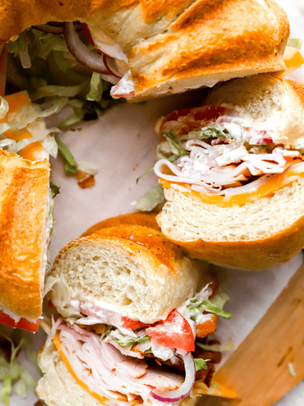 A close-up of the finished party sandwich, cut into sections, highlighting the crispy bacon, melted cheese, fresh vegetables, and soft bread layers.